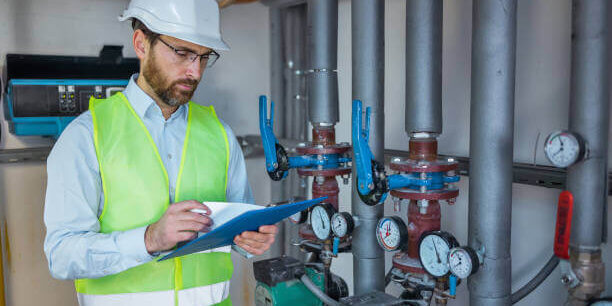 Industrial engineer standing by pipes in heating power plant and make notes in clipboard